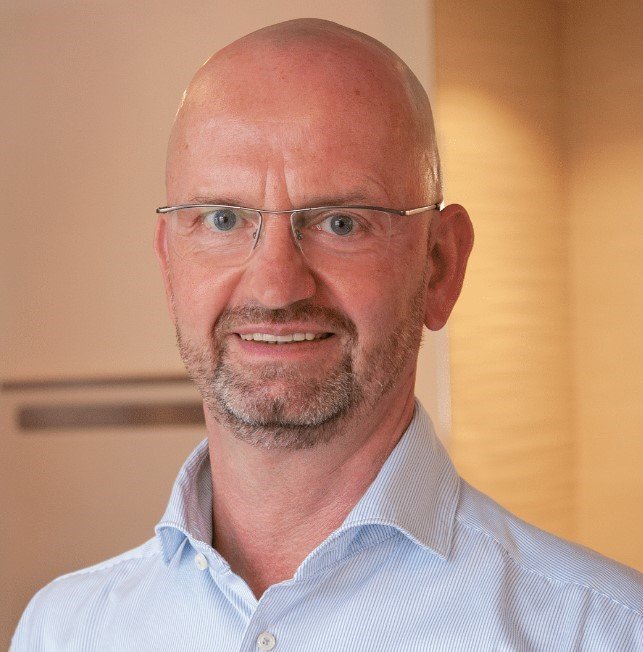 Der Mann mit Brille und Bart strahlt Freundlichkeit aus, während er in einem hellen, stilvollen Raum posiert.