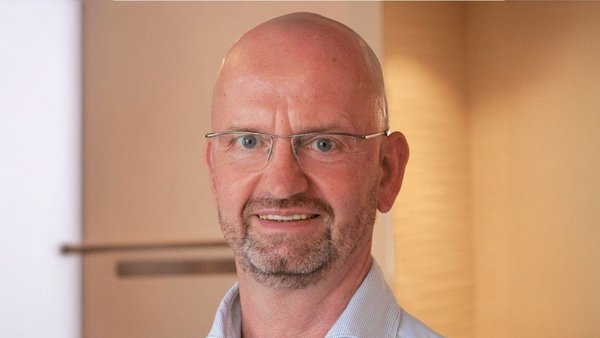 Ein lächelnder Mann mit Glatze und Brille steht vor einem hellen, neutralen Hintergrund und strahlt Freundlichkeit aus.